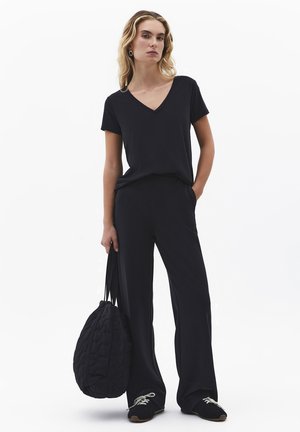 Frau trägt ein schwarzes V-Ausschnitt-Oberteil und weit geschnittene Hose, hält eine große schwarze gesteppte Tasche und steht vor weißem Hintergrund.