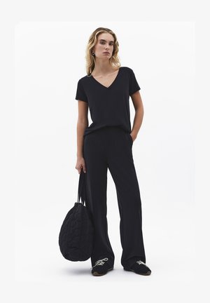 Femme portant un haut noir à col en V et un pantalon large, tenant un grand sac matelassé noir, se tenant devant un fond blanc.