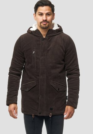 Hombre con una chaqueta con capucha de pana marrón, forrada de felpa blanca, con detalles de cremallera y botones, de pie frente a un fondo liso.