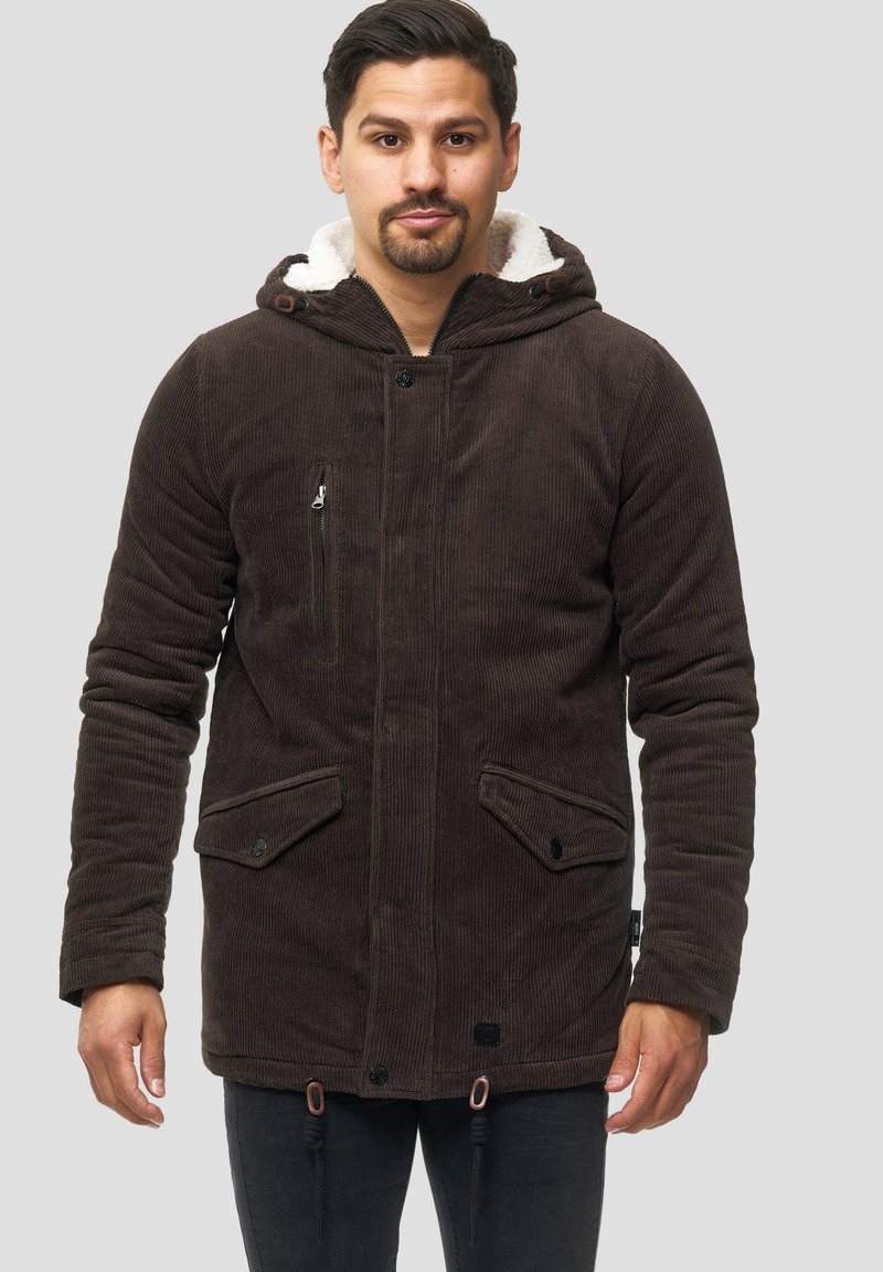 Muškarac nosi smeđu kordura hooded jaknu s bijelim krznom, sa detaljima na zatvaraču i gumbima, stoji naslonjen na neutralnu pozadinu.