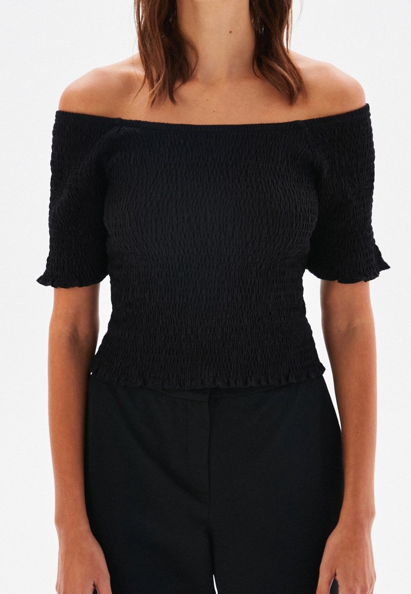 Femme portant un haut noir smocké échancré à épaules dénudées avec des manches courtes et un pantalon noir sur un fond blanc uni.