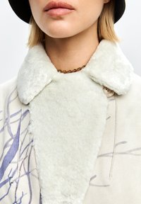 Primo piano di una persona che indossa un cappotto con colletto in pelliccia sintetica bianca, una collana con catena dorata e un cappello nero.