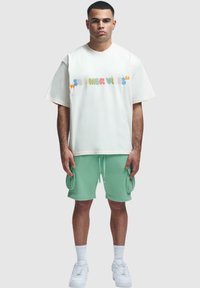Weißes oversized T-Shirt mit mehrfarbigem "SUMMER VIBES"-Druck, kombiniert mit mintgrünen Cargo-Shorts und weißen Sneakers. Aus weichem Baumwollmix.