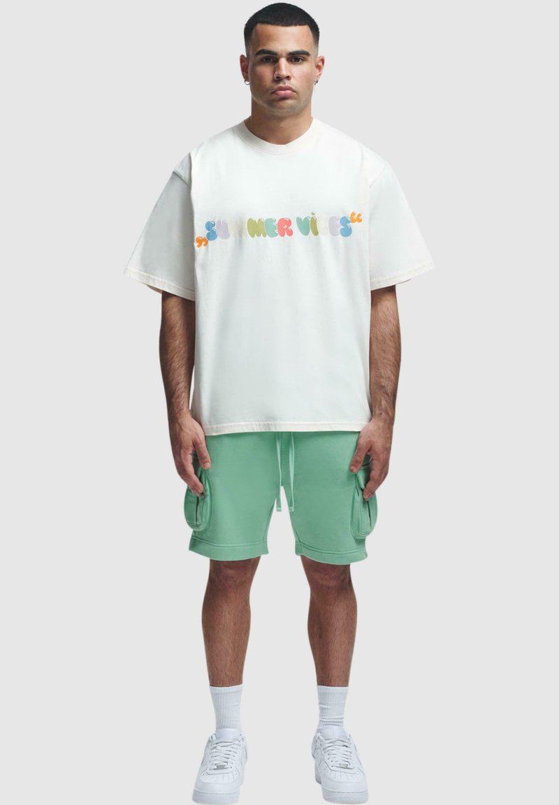 Weißes oversized T-Shirt mit mehrfarbigem "SUMMER VIBES"-Druck, kombiniert mit mintgrünen Cargo-Shorts und weißen Sneakers. Aus weichem Baumwollmix.