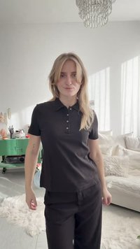 Jeune femme aux cheveux blonds portant un polo noir et un pantalon, debout dans un salon lumineux avec un meuble vert et un canapé blanc.