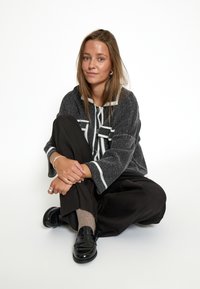 Sivi teksturirani pulover s bijelim naglascima, oversized kroja, crne široke hlače i sjajne crne loaferice. Pozicija sjedenja na bijeloj pozadini.