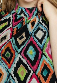 Mångfärgad tunika med ett diamantmönster i livfulla nyanser av rosa, grönt, blått, orange och svart, tillverkad av ett slätt och strukturerat tyg.