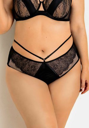 Kobieta o krągłych kształtach w czarnym koronkowym biustonoszu i dopasowanych majtkach z wysokim stanem z przezroczystymi panelami bocznymi i ozdobnym paskowym detalem na jasnym tle.