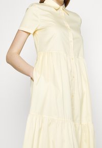 Femme portant une robe jaune pâle à manches courtes boutonnée avec un col et une jupe à volants froncée, main placée dans la poche latérale.