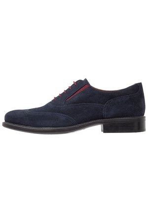 Chaussure habillée pour homme en daim bleu marine avec œillets rouges pour les lacets, détails brogue et un petit talon empilé noir.