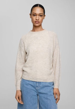 Žena s tmavými vlasy v béžovém chlupatém svetru a světle modrých džínách stojí před jednobarevným šedým pozadím.