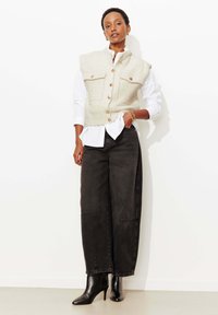 Veston en tweed crème avec des boutons dorés, porté sur une chemise blanche. Jean noir à jambes larges et bottines noires à bout pointu complètent le look.