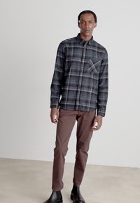 Chemise à bouton en tartan bleu foncé, manches longues, poche poitrine. Associée avec un pantalon slim marron et des bottines noires, sur un fond neutre.