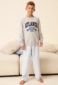 Szary sweatshirt z granatowym napisem "ATLANTA TRACK & FIELD", noszony z jasnoniebieskimi, paskowanymi spodniami od piżamy, stojąc boso na miękkiej, neutralnej kanapie.