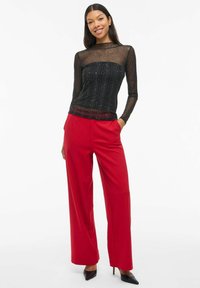Top negro de malla de manga larga con rayas brillantes, combinado con pantalones de pierna ancha rojos de cintura alta. Unos tacones negros completan el atuendo.