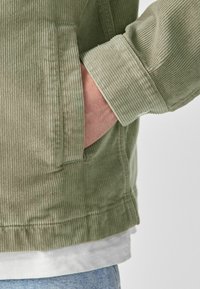 Personne portant une veste en velours côtelé vert clair avec la main insérée dans la poche latérale, une chemise blanche en dessous, et un jean bleu clair visible.
