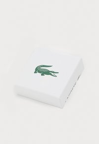 Baltas dėžutė su tekstūruota paviršiumi, kuriame viršuje iškilęs žalias krokodilo logotipas, o šone žalios spalvos tekstu išspausdintas pavadinimas "Lacoste".