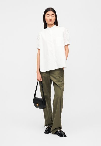 Άσπρο πουκάμισο με κοντά μανίκια και κουμπιά, με όρθιο γιακά, συνδυασμένο με ελαφρώς φαρδιά χακί παντελόνια, κρατώντας μια μικρή μαύρη τσάντα ώμου.