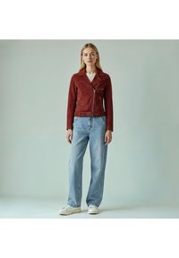 Rød ruskind bikerjakke med bælte i taljen, lynlåslommer og hakket krave, kombineret med lyseblå viddebukser og hvide sneakers.