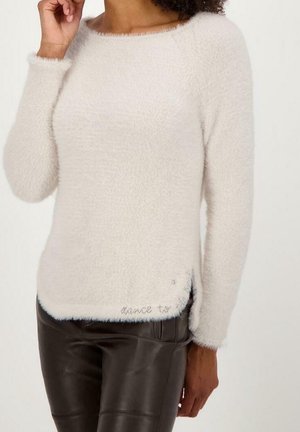 Vrouw draagt een zachte, crèmekleurige donzige trui met lange mouwen en een zwarte leren broek, poserend met één hand bij haar gezicht.