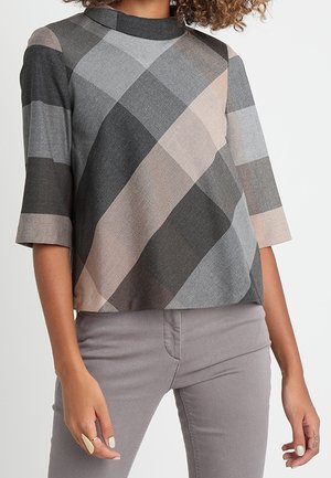 Femme portant un haut à motif géométrique gris, beige et noir avec des manches trois-quarts et un pantalon gris clair.