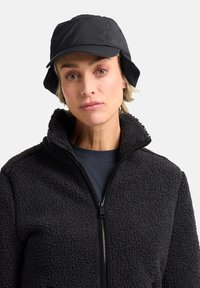 Cappello a secchiello nero con una larga visiera e un logo sottile; abbinato a una giacca in pile nera con zip e texture.