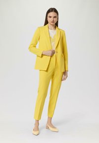 Completo giallo su misura con blazer a un bottone e pantaloni affusolati. La modella indossa una maglietta bianca con motivo giallo e tacchi beige.