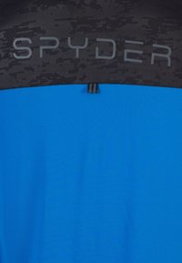 Camiseta atlética azul con panel superior negro texturizado. Presenta el logo "SPYDER" en el pecho y una pequeña etiqueta negra en el dobladillo. Textura suave.