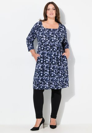 Robe florale marine à manches trois-quarts, taille cintrée, poches latérales et jupe évasée. Associée à des leggings noirs et des talons.