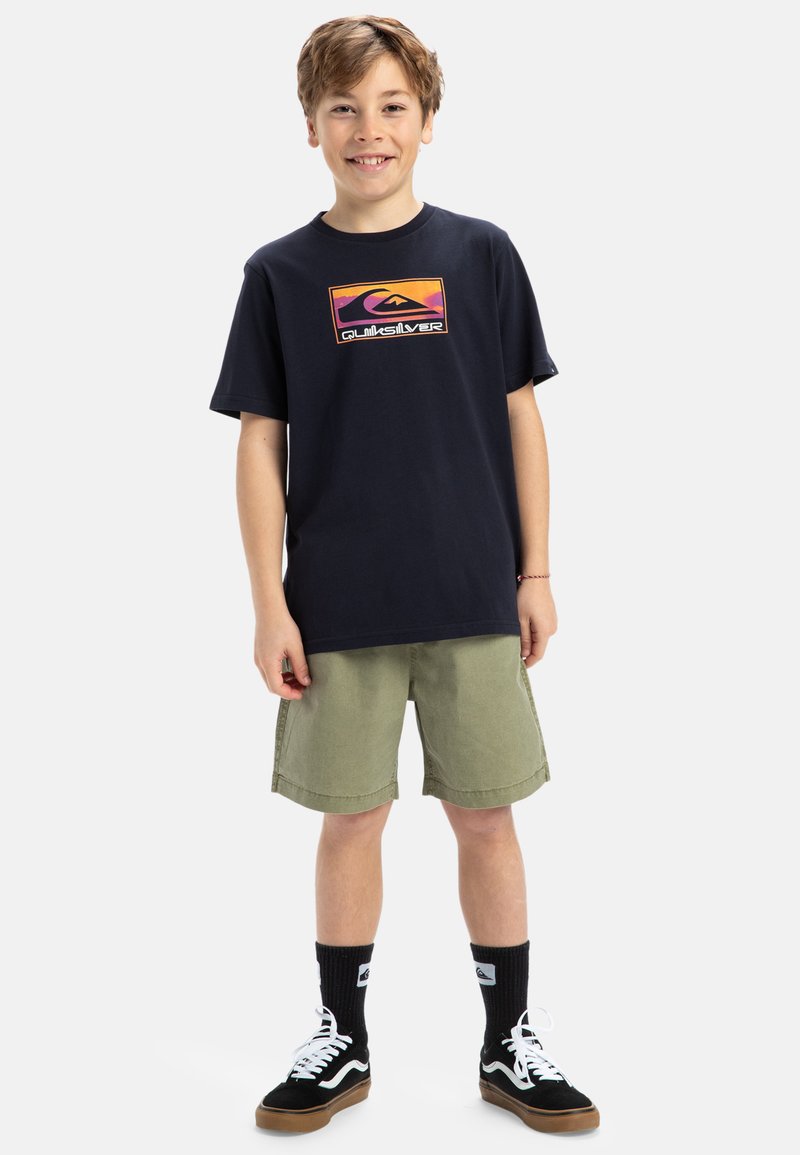 Marineblaues Baumwoll-T-Shirt mit einem bunten Quiksilver-Logo, kombiniert mit olivgrünen Shorts. Schwarze Turnschuhe mit weißen Schnürsenkeln und schwarzen Socken.