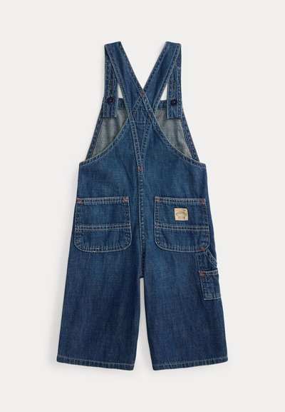 Jeans overall v temno modri barvi z nastavljivimi križnimi naramnicami, dvema sprednjima žepoma in stranskim žepom z oznako. Šivi v kontrastni rdeči barvi.