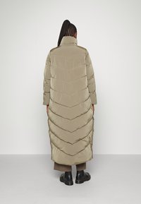 Abrigo de plumas largo color caqui con cuello alto, con costuras en forma de chevrón, bolsillos laterales y un corte relajado, combinado con botas negras gruesas.