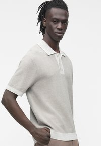 Mladý muž s dredy, který nosí světle šedé texturované polo tričko s krátkým rukávem, límečkem a légou na knoflíky, stojící s rukou v kapse.