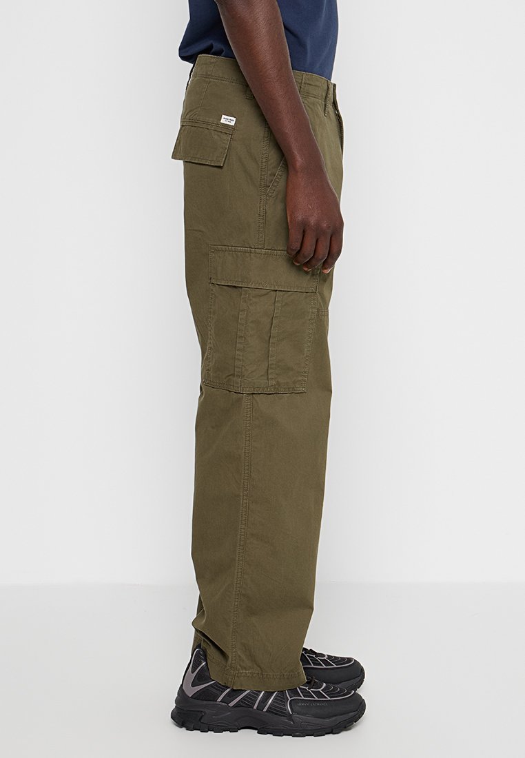 Eine Person trägt olivgrüne Cargo-Hosen mit seitlichen Taschen und schwarze Sportschuhe, steht vor einem weißen Hintergrund.