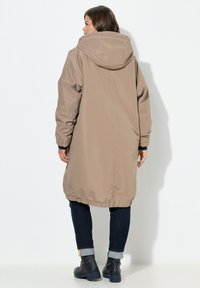 Beige langer Mantel mit Kapuze, elastischem Saum und weiten Ärmeln. Über dunklen Jeans mit umgeschlagenen Säumen und Ankle Boots für einen lässigen Look getragen.