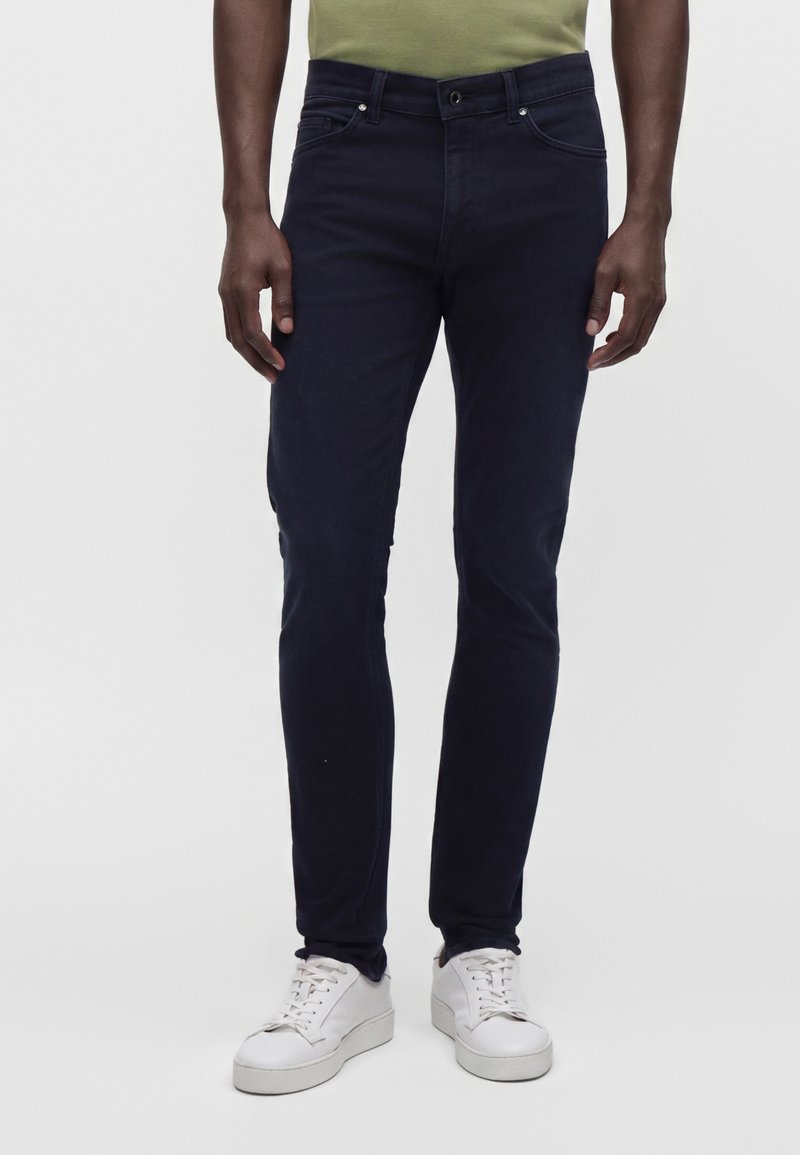 Άνδρας που φοράει σκούρο μπλε τζιν slim-fit, λευκά αθλητικά παπούτσια και ανοιχτοπράσινο πουκάμισο, στέκεται μπροστά σε ένα απλό φωτεινό φόντο.