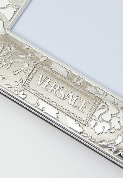 Srebrna metalowa ramka na zdjęcia z misternymi kwiatowymi wzorami oraz centralnym prostokątnym akcentem z wytłoczonym logo "VERSACE".