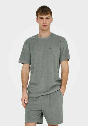 Young man wearing matching gray terry cloth short-sleeve shirt and shorts, standing with one hand in pocket against plain background.