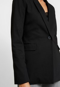 Blazer nero con una texture liscia, chiusura a un bottone, revers a punta e tasca laterale, design sartoriale, silhouette aderente.