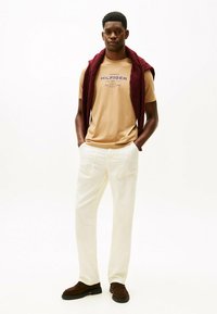 Beige bomulds t-shirt med blåt logo, cremede bukser og brune ruskinds slip-on sko. Mørkerød trøje draperet over skuldrene.