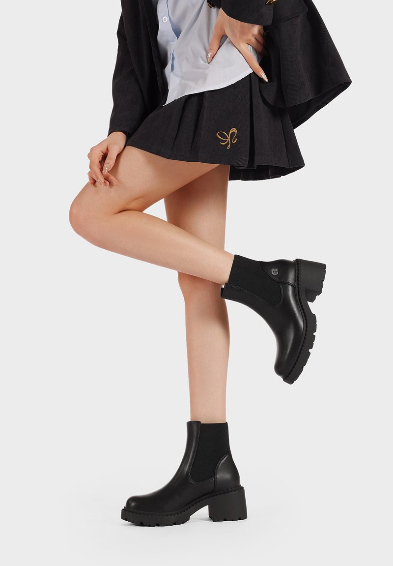 Gambe che indossano stivaletti neri con tacco spesso e una gonna plissettata nera con un simbolo ricamato in oro sul davanti.