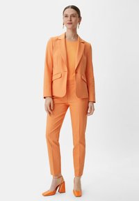 Pomarańczowa dopasowana marynarka i pasujące spodnie, wykonane z gładkiego materiału. Posiadają slim fit z klapami, zestawione z pomarańczowymi butami na słupku.