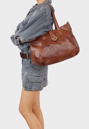 Persona che indossa giacca di jeans e shorts con borsa a tracolla in pelle marrone con chiusura a anello metallico e doppi cinturini.