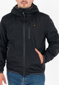 Giacca nera leggera con cappuccio, frontale con zip, tasche laterali, polsini elasticizzati e finitura testurizzata. Presenta il logo del marchio sulla spalla.