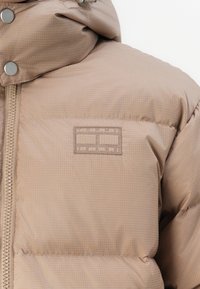 Beige dunjakke lavet af quiltet stof, med lynlåslukning og logo-patch. Hætten kan justeres med trykknapper.