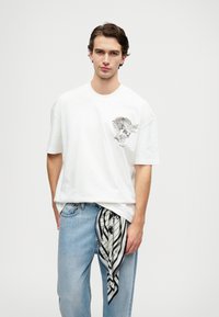 Λευκό βαμβακερό μπλουζάκι με κοντά μανίκια, που διαθέτει μαύρο και άσπρο γραφικό σχέδιο στο αριστερό στήθος. Ανοιχτό μπλε τζιν παντελόνι με μια μαύρη και άσπρη μαντήλα δεμένη στη μέση.