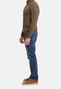 Uomo in piedi di profilo che indossa un maglione marrone con zip, jeans blu e sneakers in camoscio marrone con suole beige su uno sfondo bianco.