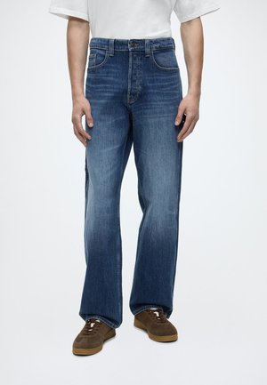 Uomo che indossa jeans blu a gamba dritta, t-shirt bianca e sneakers marroni, in piedi davanti a uno sfondo chiaro e uniforme.