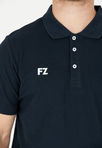 Mörkblå pikétröja i strukturerad tyg, med en treknapplåsning och vit broderad "FZ"-logotyp på vänster bröst.
