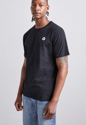 Svart bomulls t-skjorte med korte ermer, rund hals, og et hvitt logo-patch på brystet. Glatt tekstur, avslappet passform.
